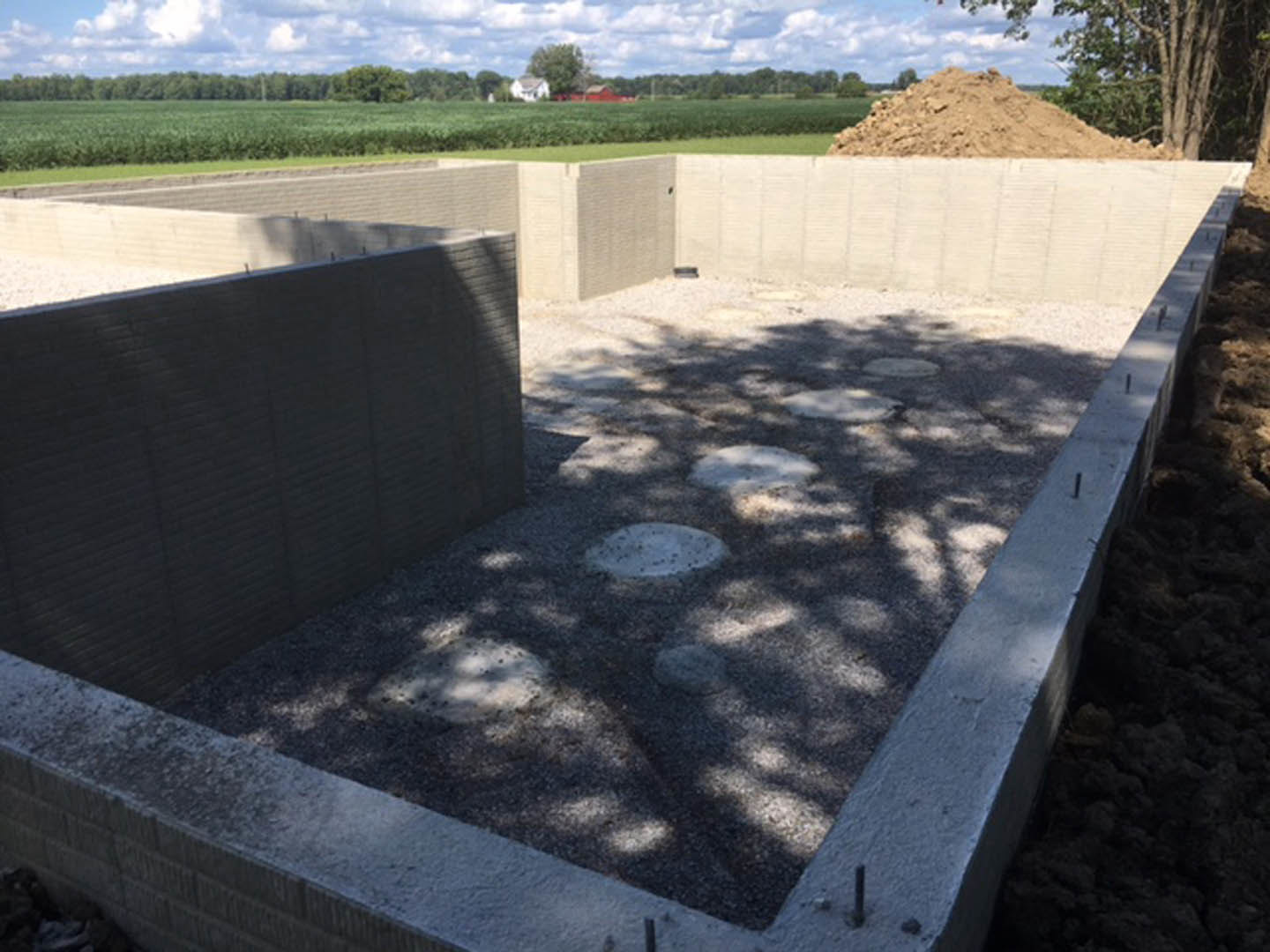 Concrete foundation slab surrounded by dirt piles, trees, and cloudy sky in the background