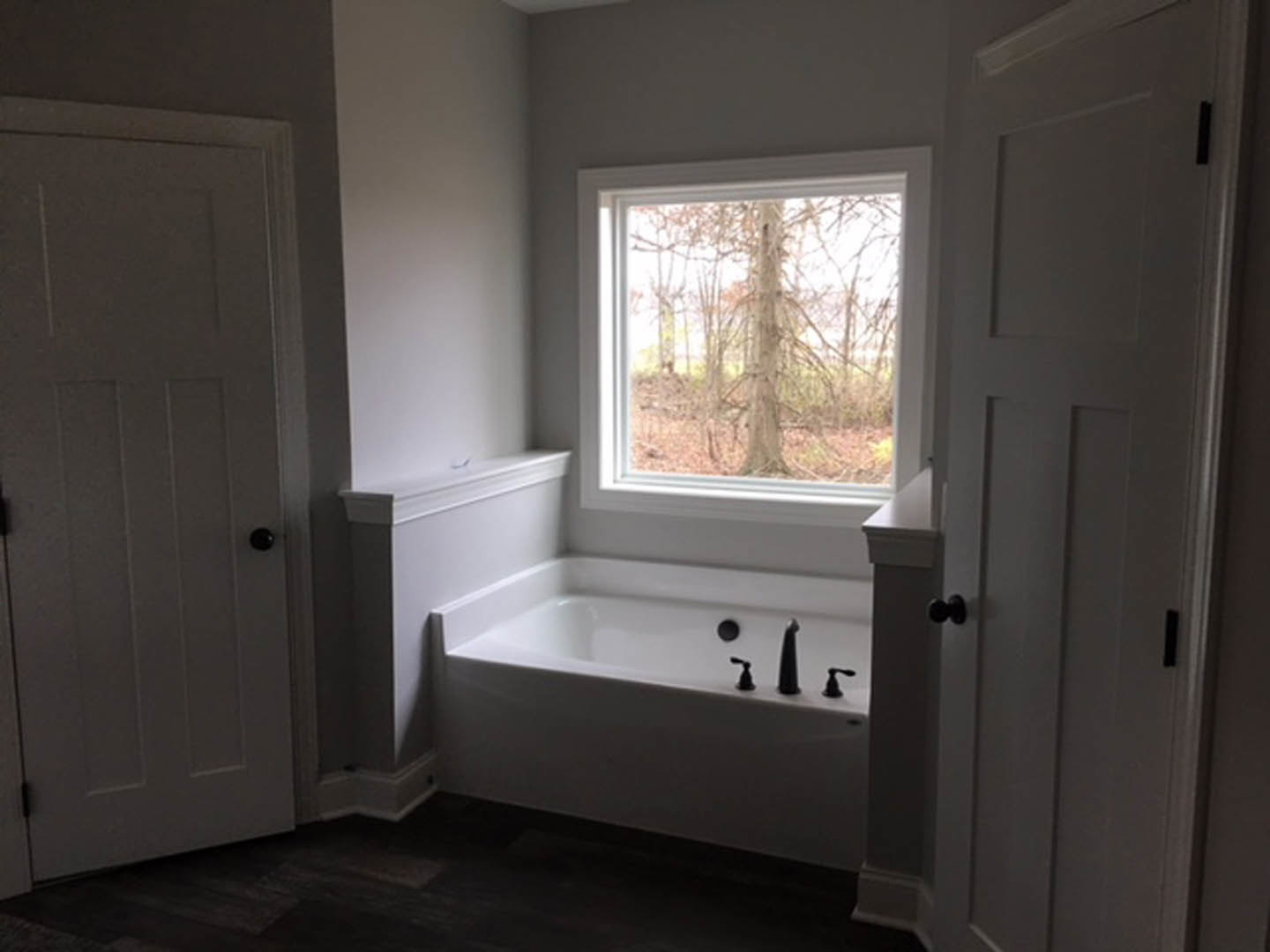 Freestanding white bathtub with chrome faucets and black knob, set beside a large window overlooking a leafy tree, light-toned walls and floor visible