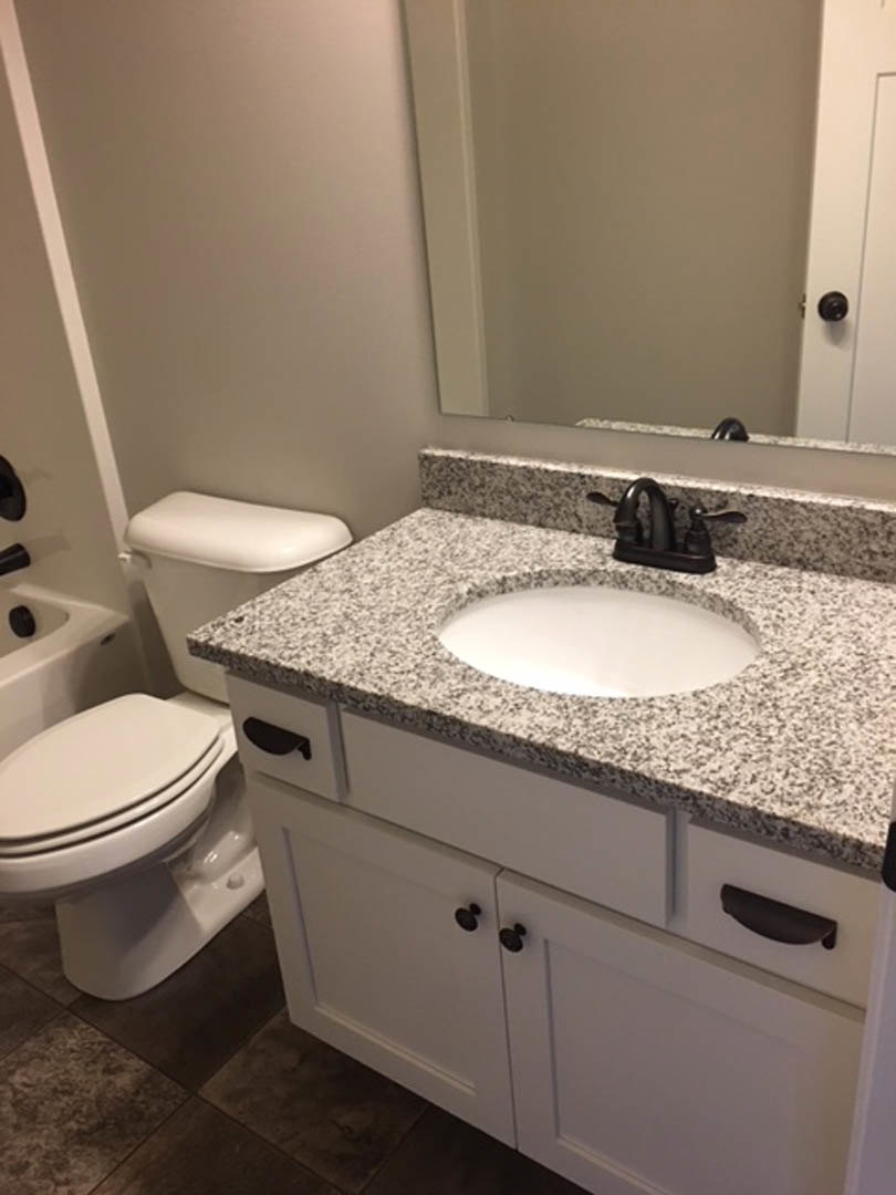 Modern bathroom with white porcelain sink, chrome faucet, rectangular mirror above, white toilet beside, and white vanity cabinet with black handles against light tiled walls