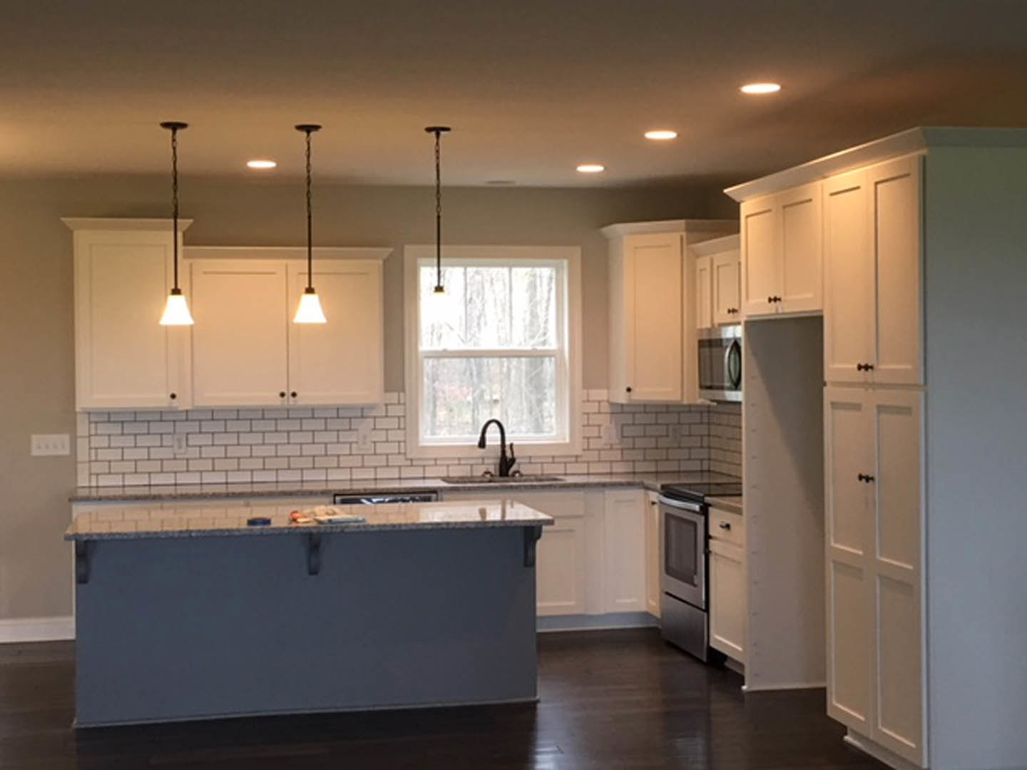 White kitchen with shaker cabinets, central island with quartz countertop, stainless steel stove, tile backsplash, large window letting in natural light, wood flooring