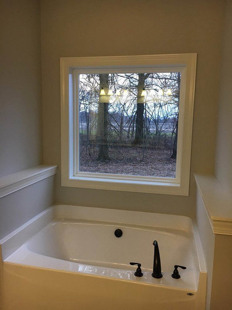 Freestanding white bathtub beneath large window with outdoor lights visible, black faucet and handle, white shelf against white wall, close-up bottle on tub ledge