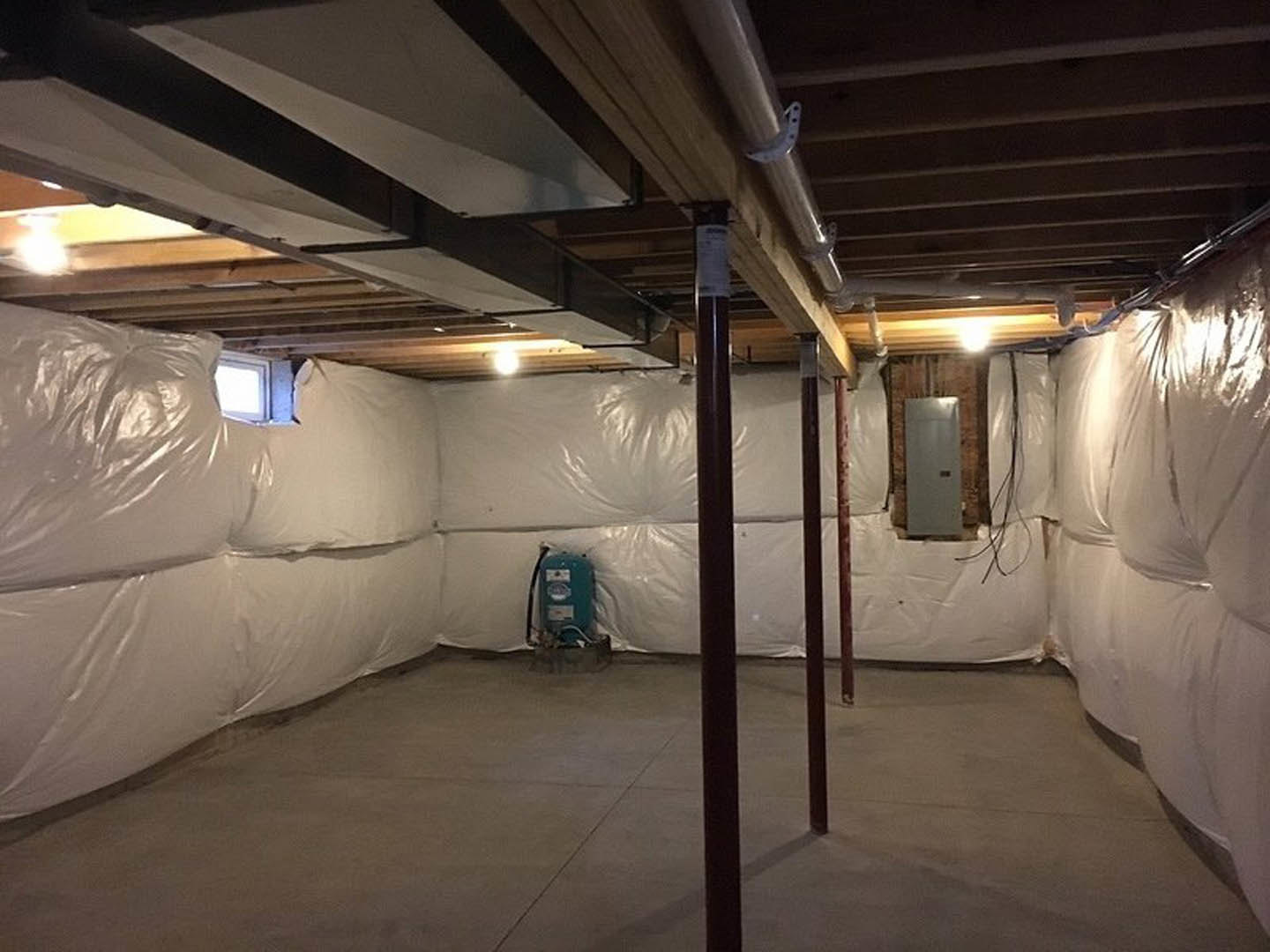 Basement with concrete floor and white plastic coverings on walls and ceiling, black support pole, white door with mounted square object, close-up of machinery, scattered white