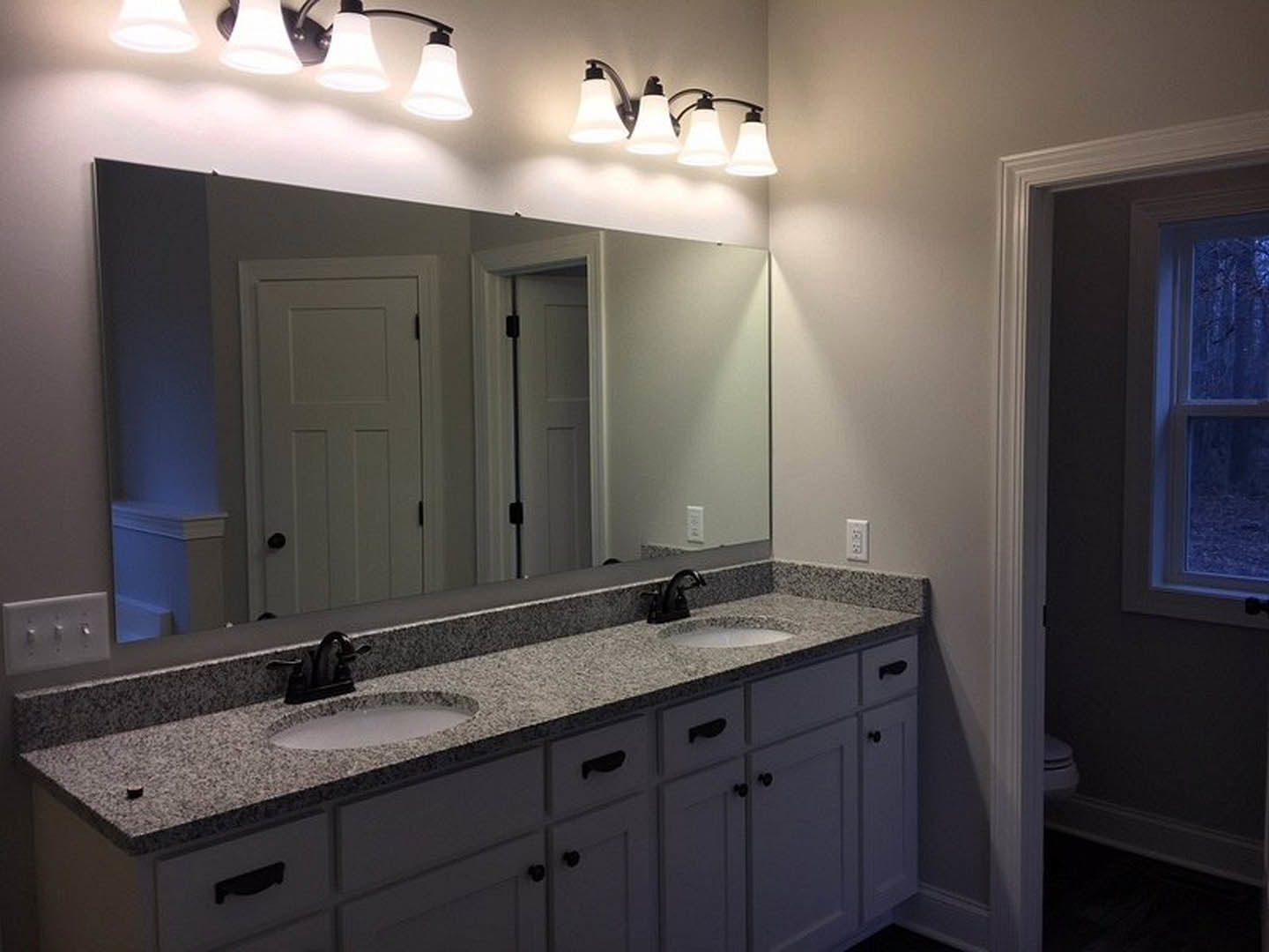 Double vanity bathroom with white countertop, black faucets, large wall mirror, two doors reflected, and modern light fixture above sinks.