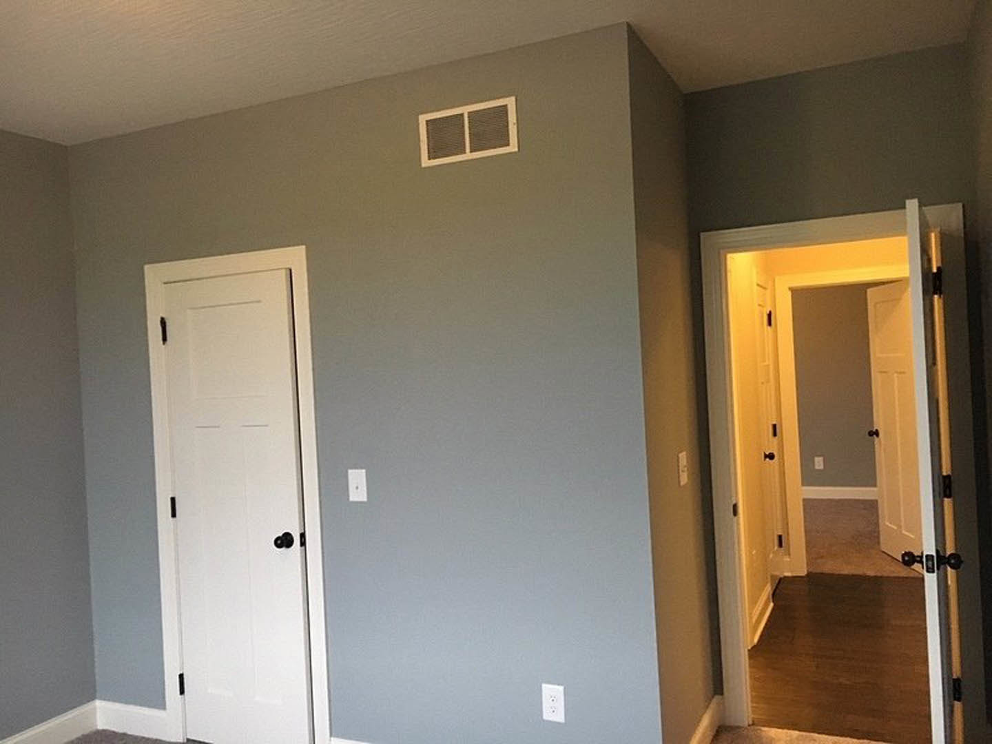 Hallway with dark wood flooring, white walls, wall vent, and white door with black knobs open to adjacent room