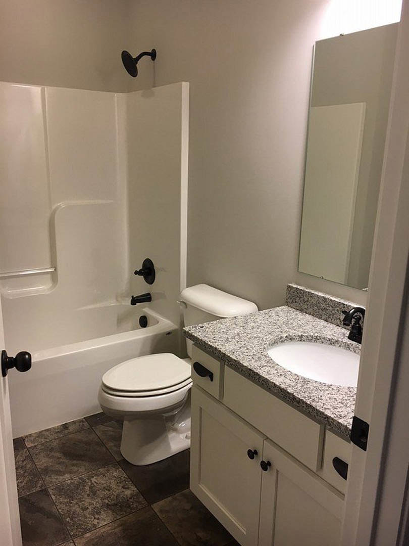 Bathroom with white porcelain sink and toilet, grey countertop, white bathtub, black wall hook, black door knob on white door, light-colored tile flooring and walls.