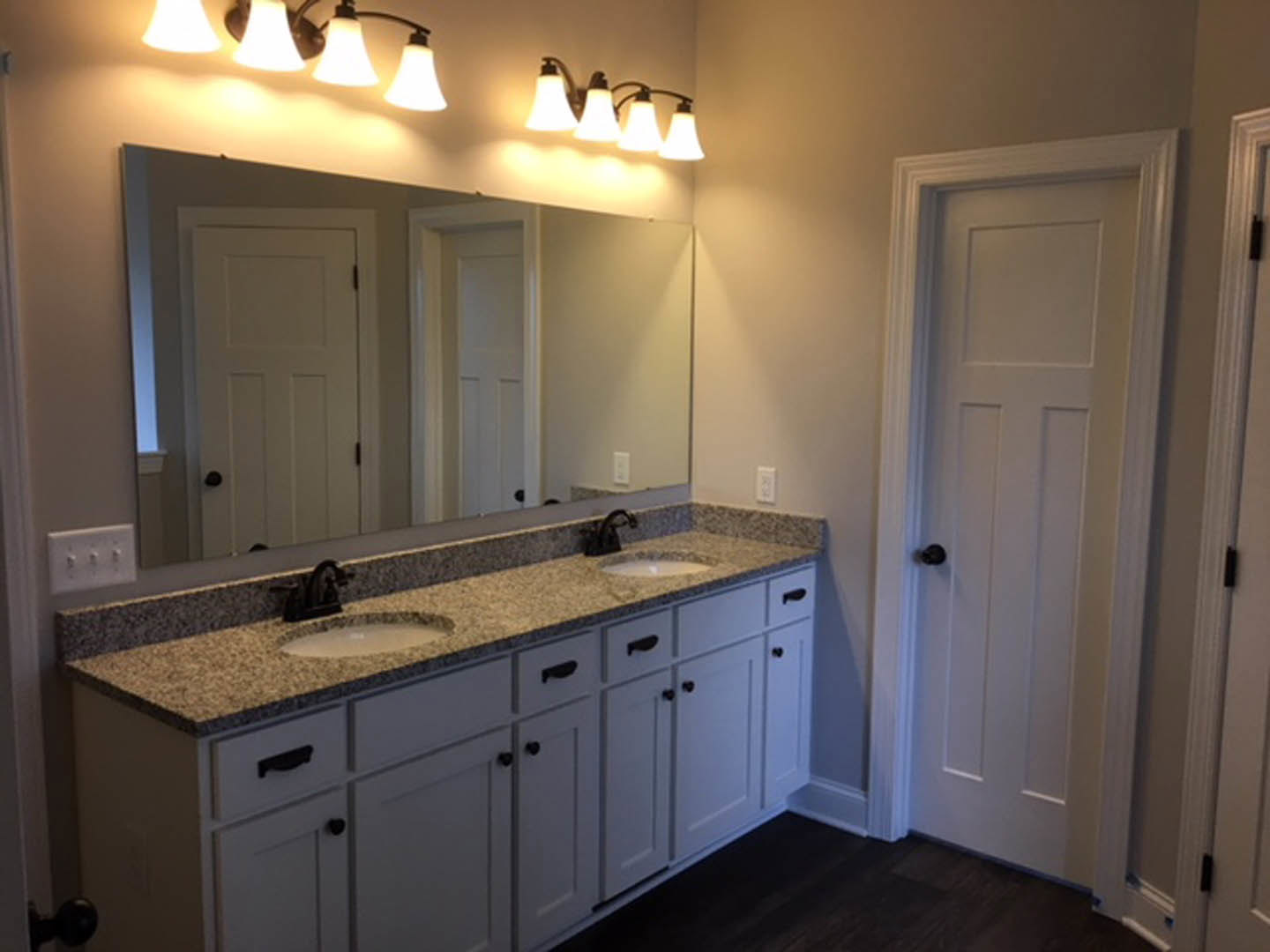 Bathroom with double vanity featuring white sinks, stone countertop, wall-mounted mirror, two-lamp light fixture, white door with black knob, and tile flooring