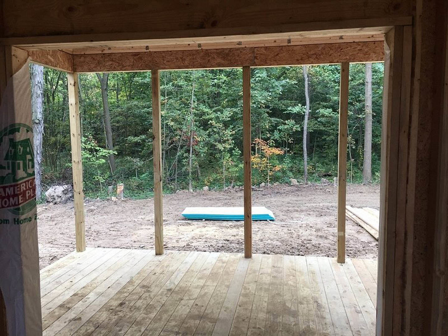 Open room with light wood flooring, blue ottoman centered, large windows framing outdoor view with trees and dirt field, wood beam visible overhead.
