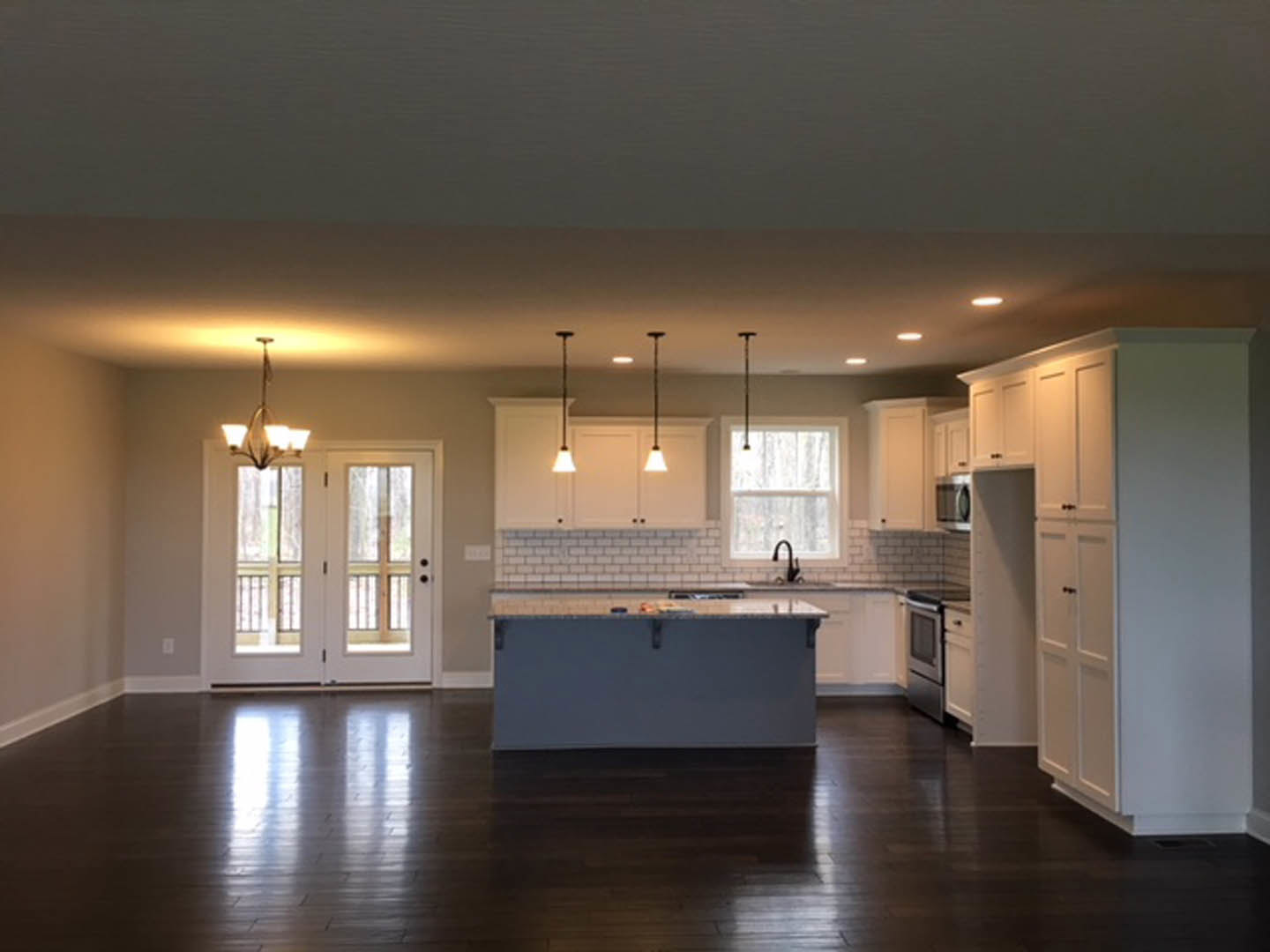 Kitchen with white shaker cabinets, large central island with stone countertop, wooden flooring, glass-paneled double doors, window above sink, pendant light fixture, white tile