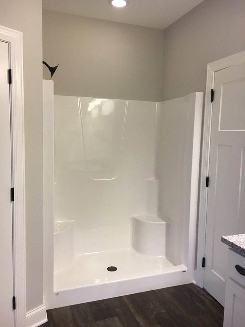White tile shower with chrome showerhead, dark wood floor, white baseboards, white door with black handle, glass lamp and glass decor visible
