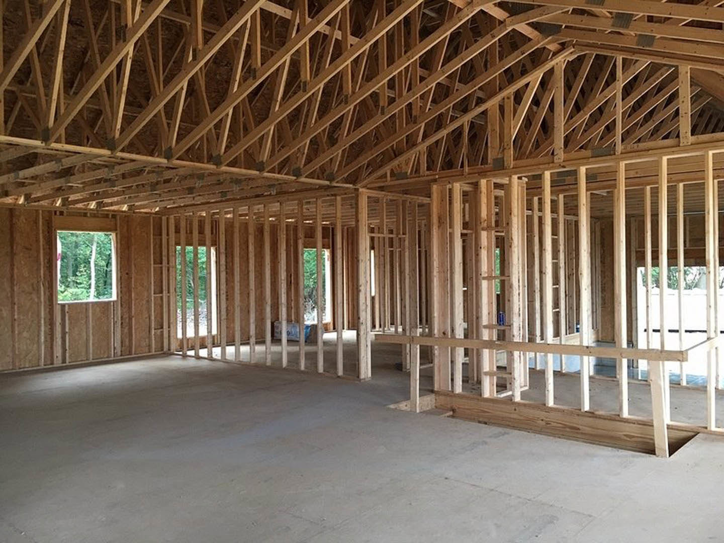 Wood-framed house under construction with exposed ceiling beams, concrete floor, white support posts, and large window overlooking trees