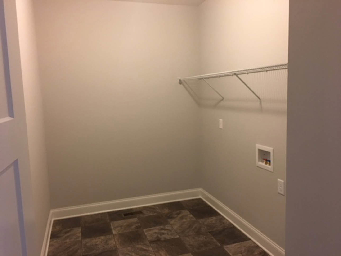 Empty room with smooth white plaster walls, light gray tile flooring, white square shelf with small hole, white metal rod mounted on wall, and white door partially visible.