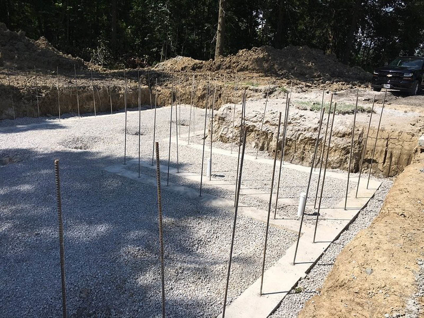Concrete house foundation with exposed metal poles, surrounded by dirt and gravel, black car parked roadside, trees and plants in the background.