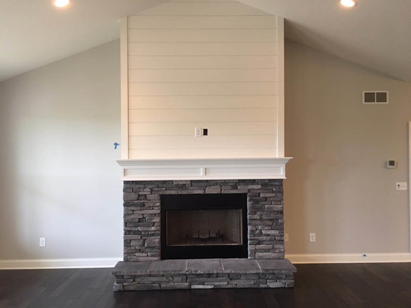 Stone fireplace with black metal frame set in white-walled living room, large square window to the side, hardwood flooring, and brick hearth border