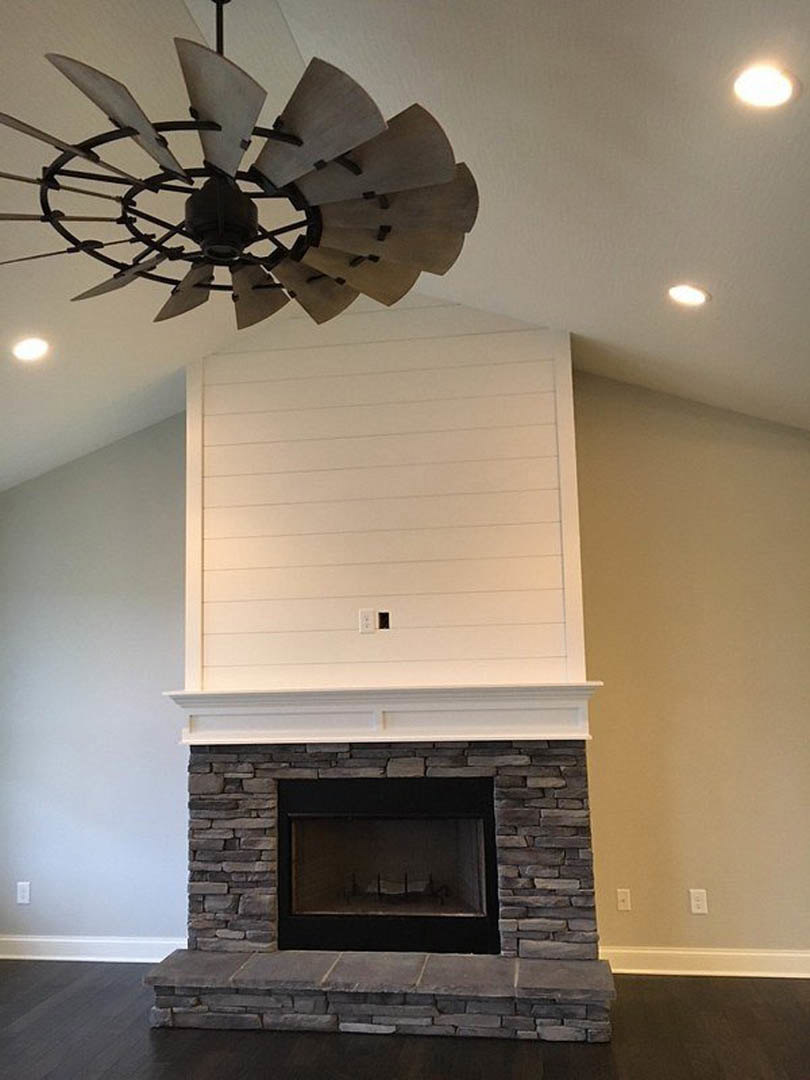 Ceiling fan with dark blades mounted above a living room featuring a black-framed fireplace, white walls, and brick accents.