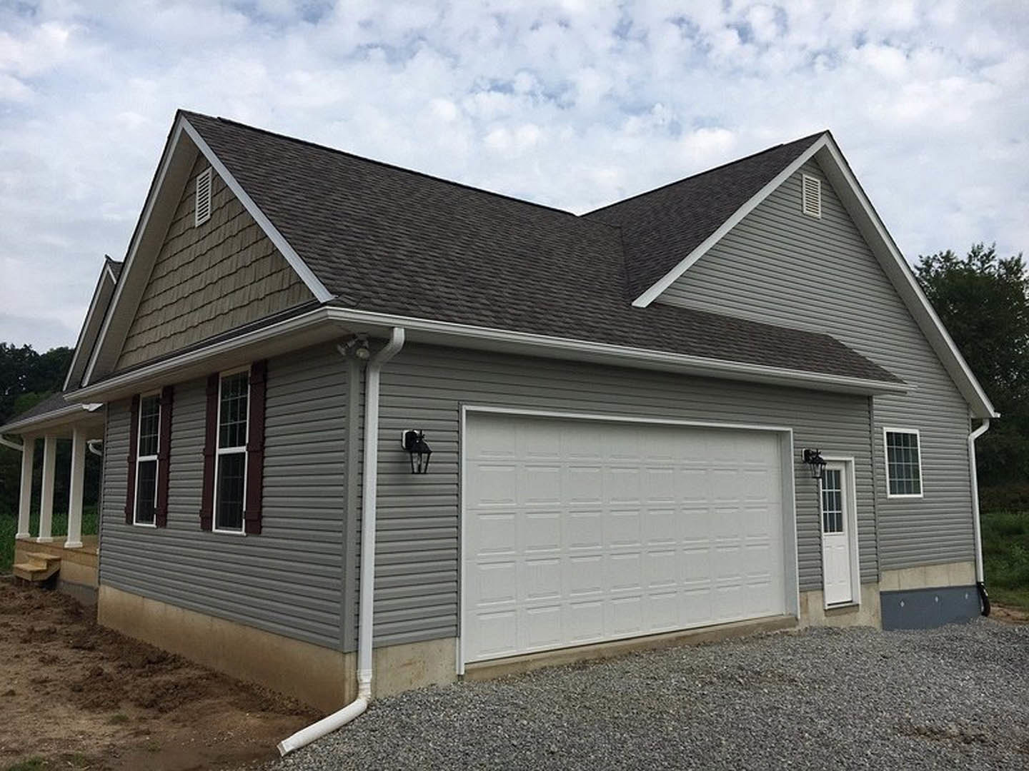 Two-story home with gray siding, white trim, attached single-car garage with paneled door, multi-pane windows, white entry door with glass panes, gravel driveway, and cloudy sky