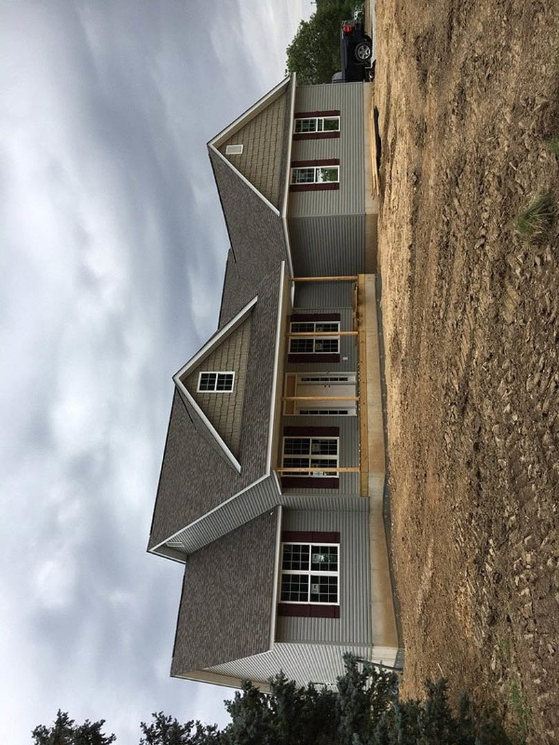 Modern house under construction featuring a wooden deck, multiple square windows, concrete retaining wall bordering a dirt field, and a tree with green leaves in the yard.