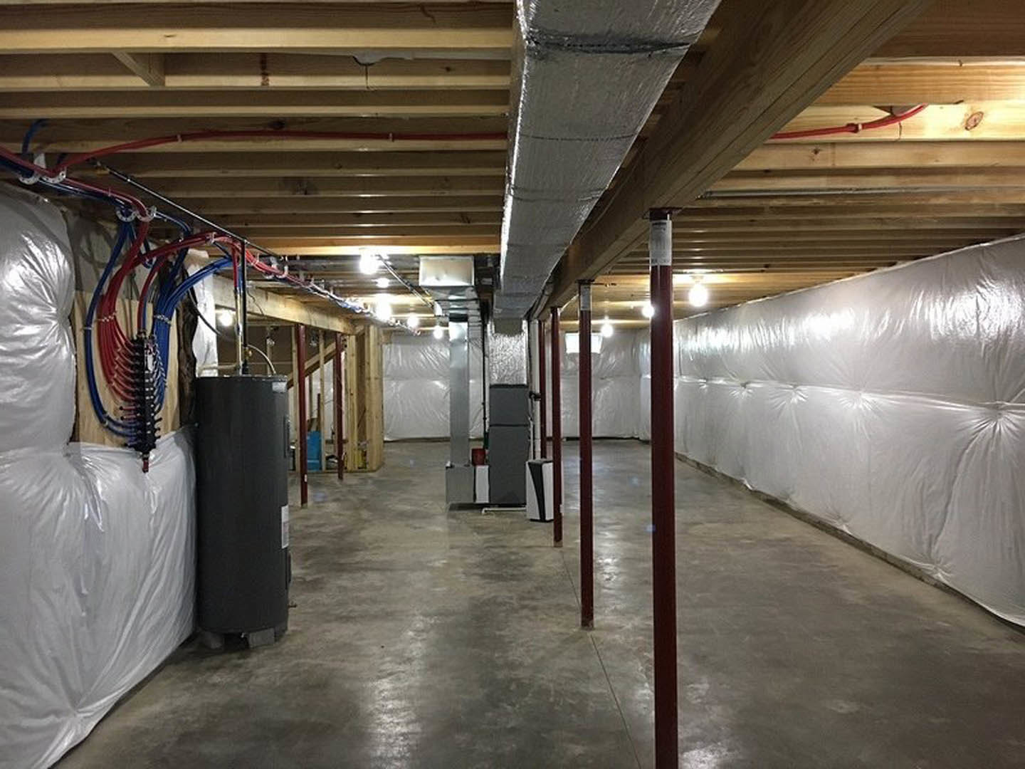 Unfinished room with white plastic-covered wall, exposed steel pipes, concrete floor with red poles, large white plastic bag with attached wires, and black cylinder with white