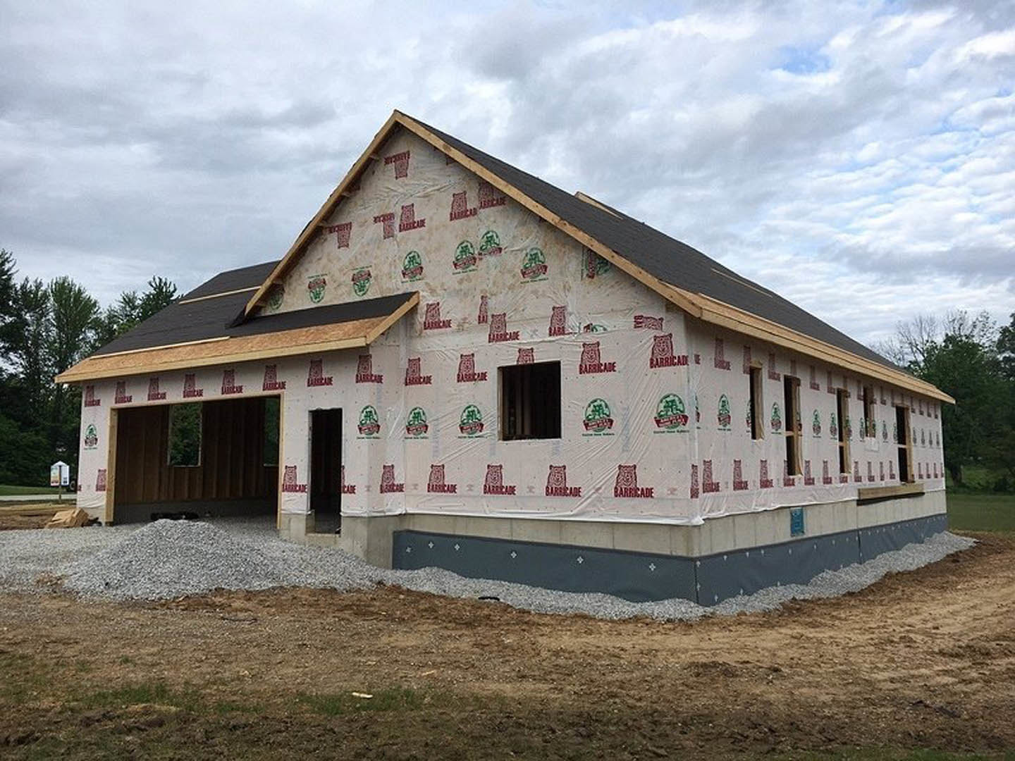 Two-story house under construction with exposed framing, covered roof, and patch of dirt in front yard; white sign with logo visible in window.