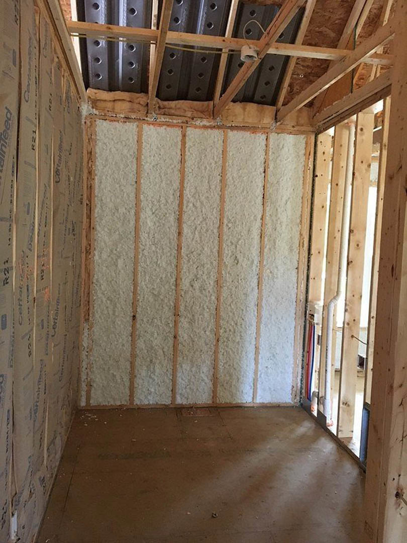 Room under construction with exposed wood beams, white foam insulation on walls, metal roof panels, and visible plumbing pipe along unfinished floor.