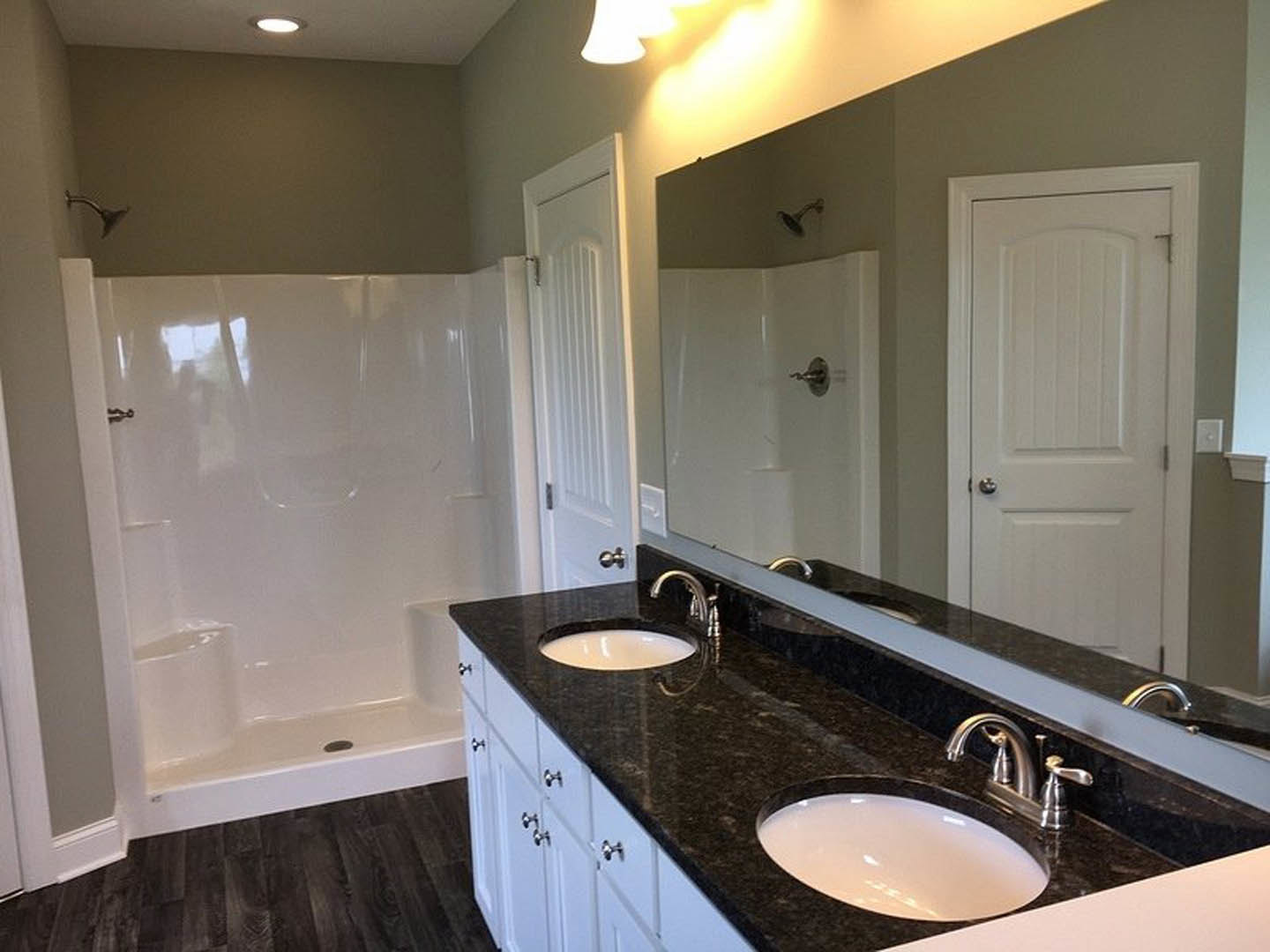 Modern bathroom featuring a wide rectangular mirror above a spacious white sink with chrome faucets, black countertop, white door with silver handle, and wall-mounted shower head.