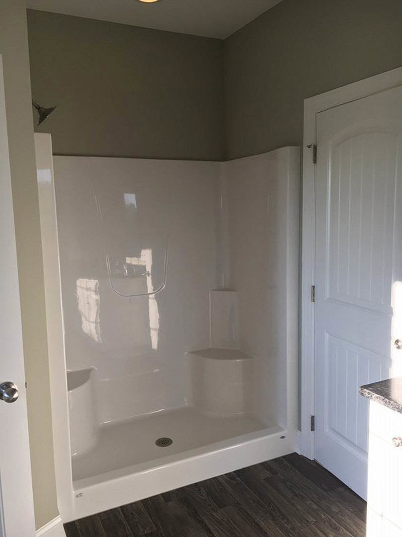 Glass shower enclosure with white tile walls, wood flooring with white trim, and a white door featuring a chrome handle