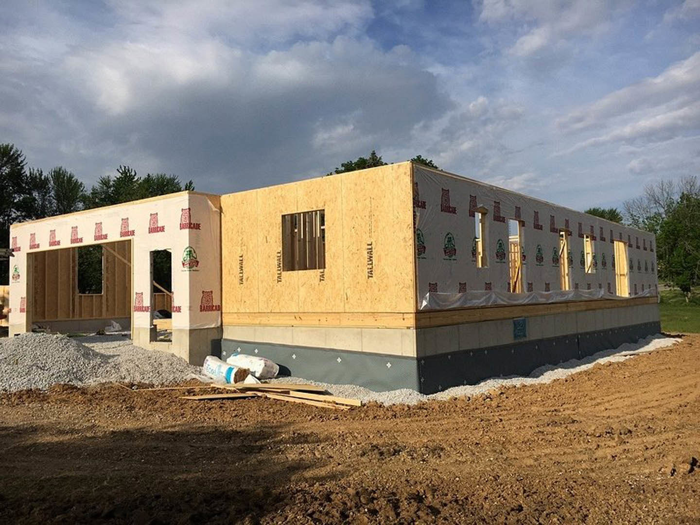 Partially built house with exposed framing, gravel piles on ground, unfinished exterior walls, single window installed, wooden fence in foreground, scattered rocks, cloudy sky