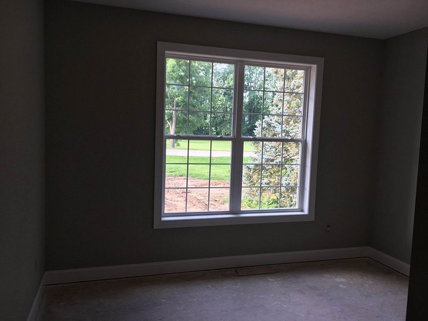 Large window with multiple square panes overlooking a dirt path and trees, grey flooring, white walls, and minimal window blinds in a bright residential room