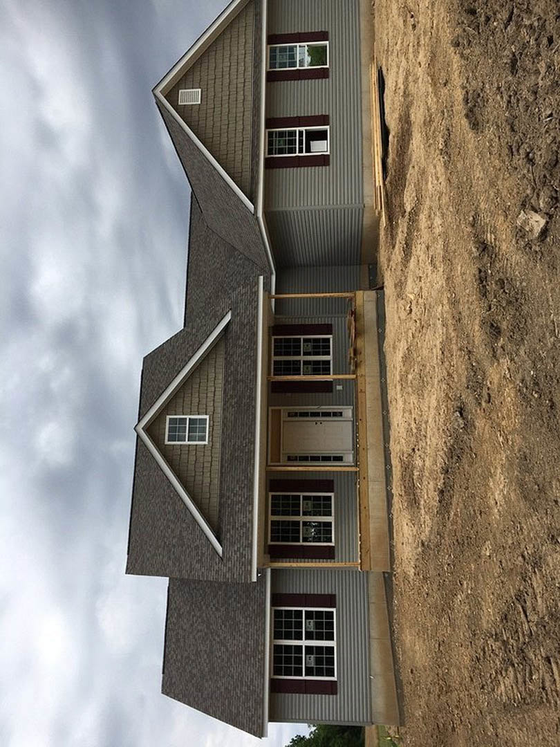 Partially built house with exposed wooden framing, dirt lot in foreground, multiple grid-style windows, white facade, cloudy sky overhead