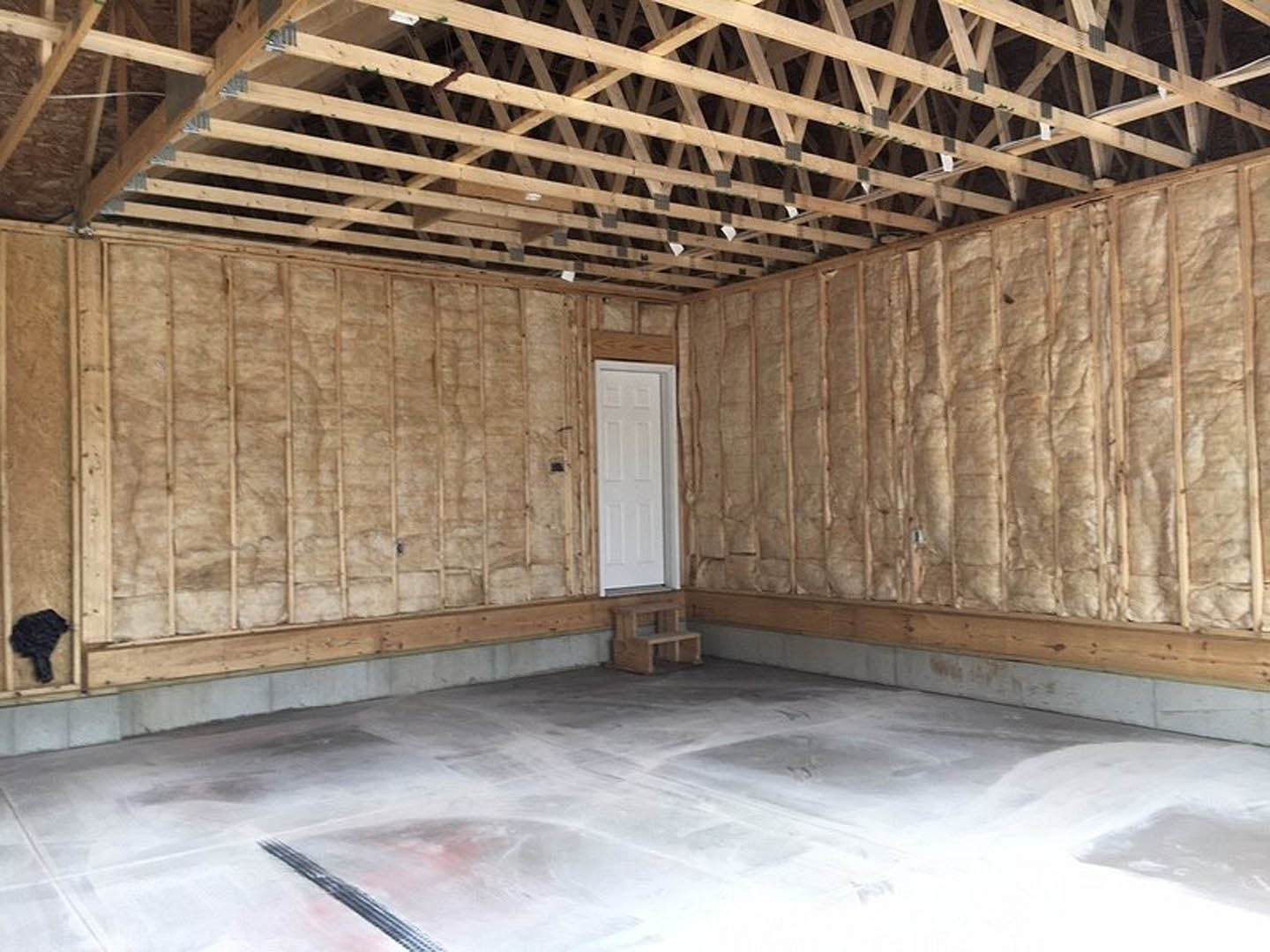 Concrete floor with close-up texture, white door framed in wood, exposed wooden roof beams, partial view of a wooden bench, neutral walls, and blurred black object in foreground