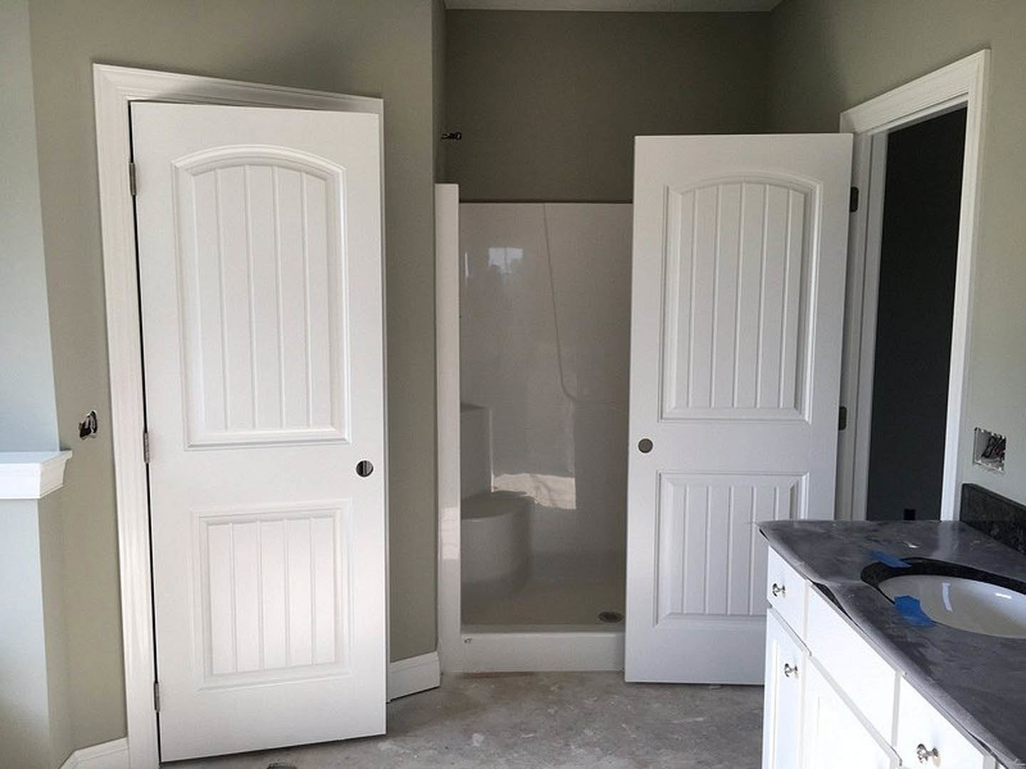 Bathroom with two white doors, glass shower enclosure, white toilet, sink with blue painter’s tape, white walls, and black-bordered countertop.
