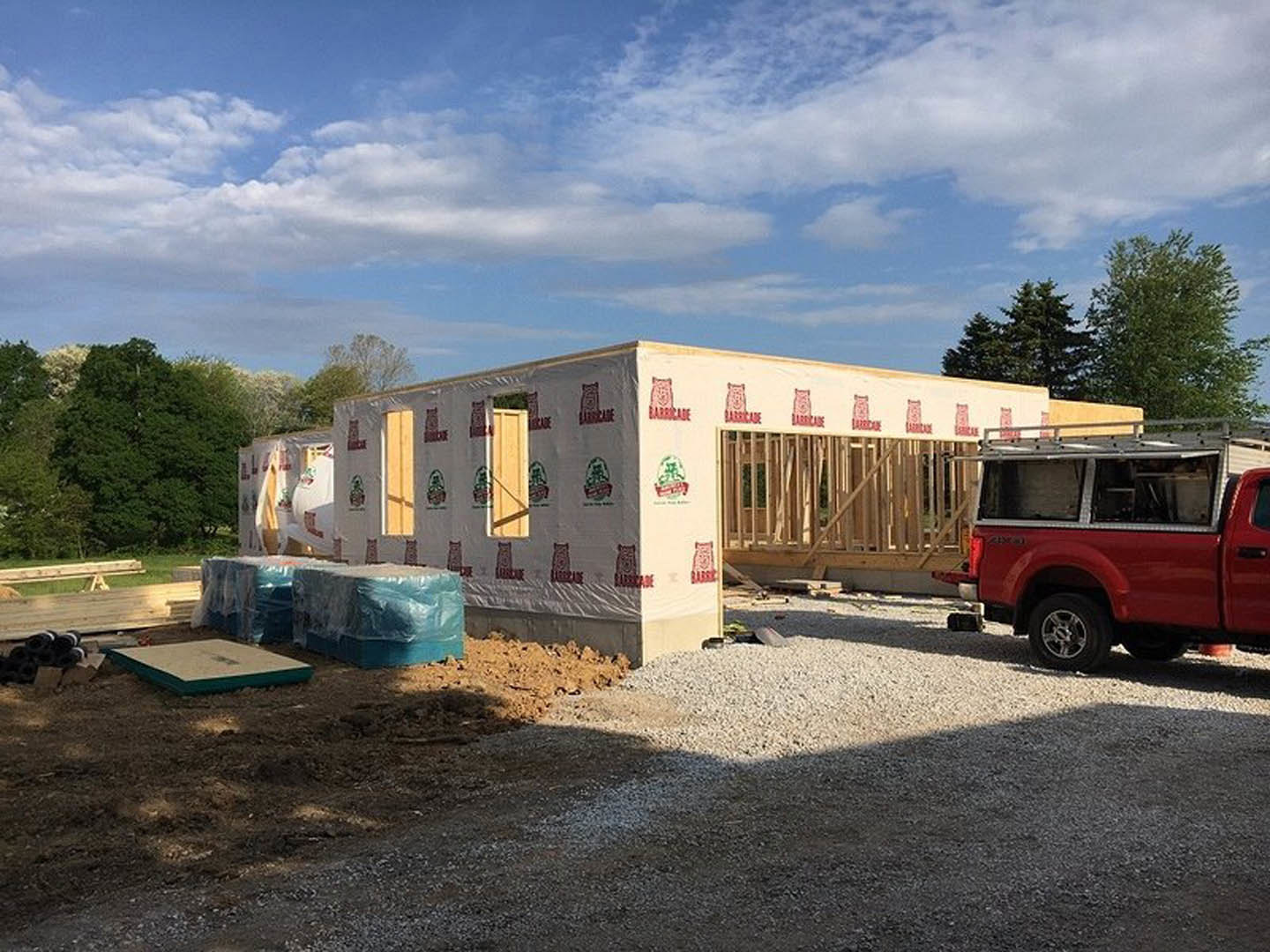 Red work truck with ladder parked beside partially constructed home with white exterior wall, blue plastic wrap, and leafy tree under a cloudy sky.