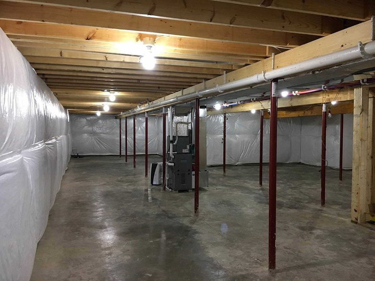 Basement with white plastic sheeting covering walls and floor, exposed ceiling beams, concrete floor, visible pipes, and a red support pole