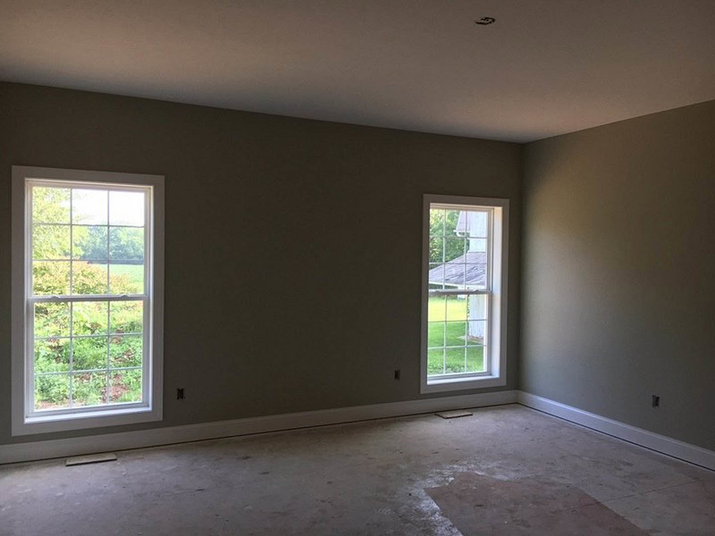 Spacious room featuring large windows with views of green grass and trees, smooth concrete flooring, white plaster walls, and natural daylight illuminating the interior.