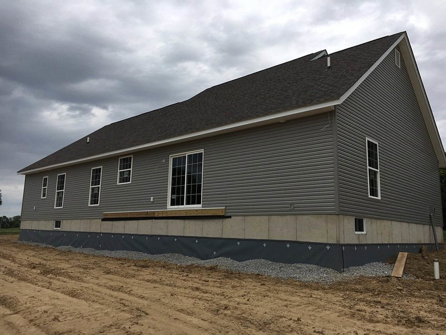 Partially built house with grey siding, white-framed window, exposed wood, dirt and gravel foreground, cloudy sky in background