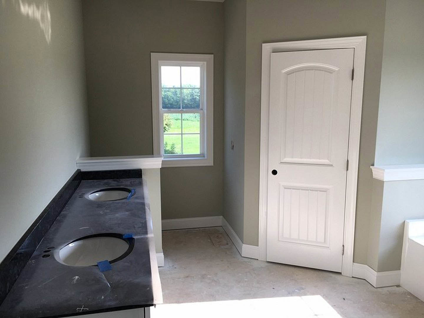 Bathroom with dual sinks set in a white countertop, white door with black handle, wall-mounted mirror, light tile flooring, and window offering outdoor view.