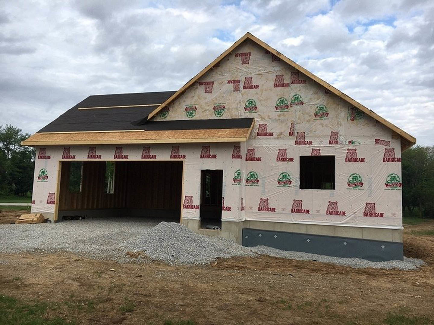 Modern custom home under construction featuring unfinished siding, exposed framing, attached garage, black square window with red bear logo, white exterior surfaces, and grassy lot