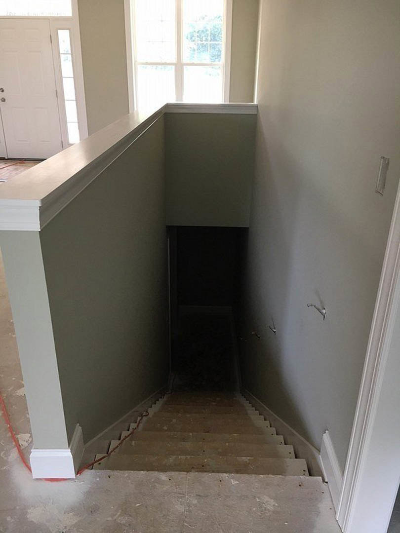 Wood staircase with white risers and handrail ascending beside a large window, sunlight streaming onto light-colored plaster walls and hardwood floor, white door with glass panel