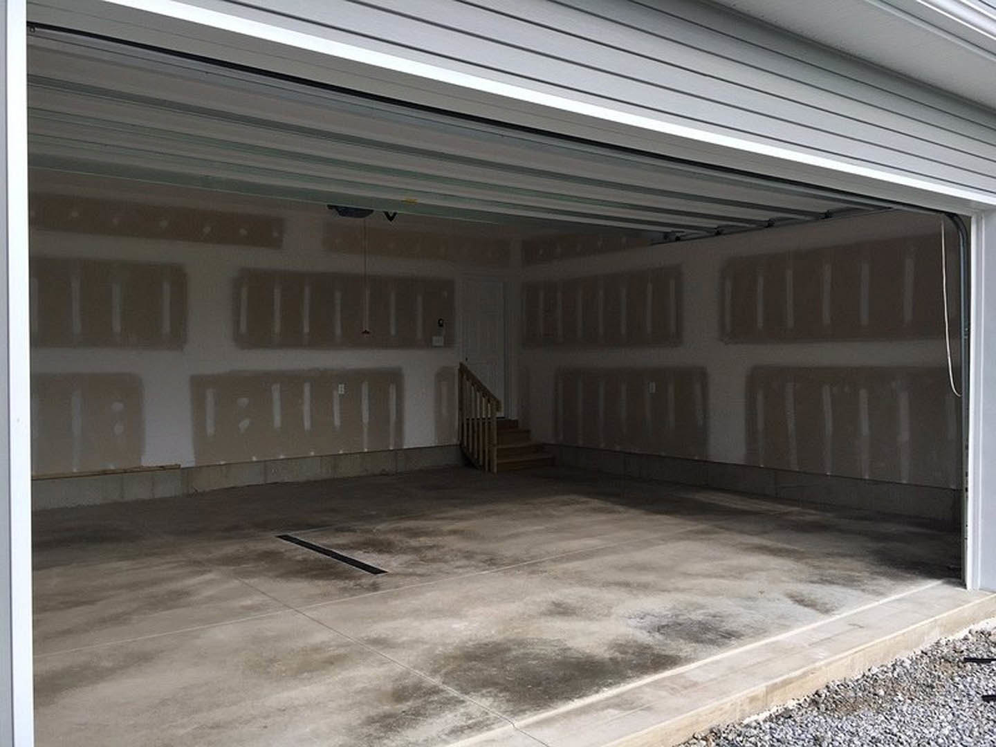Garage interior with concrete floor featuring a black line, wooden staircase leading to upper level, white walls, and ceiling-mounted light fixture