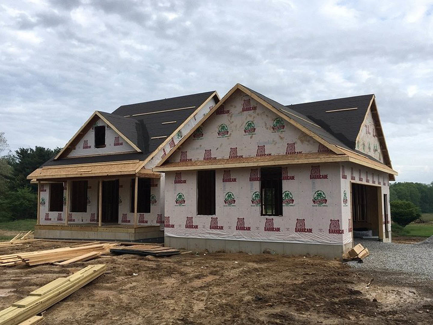 Framed house under construction with exposed wood beams, partially installed windows, pile of wood planks in foreground, graffiti on window, black curtain with white border
