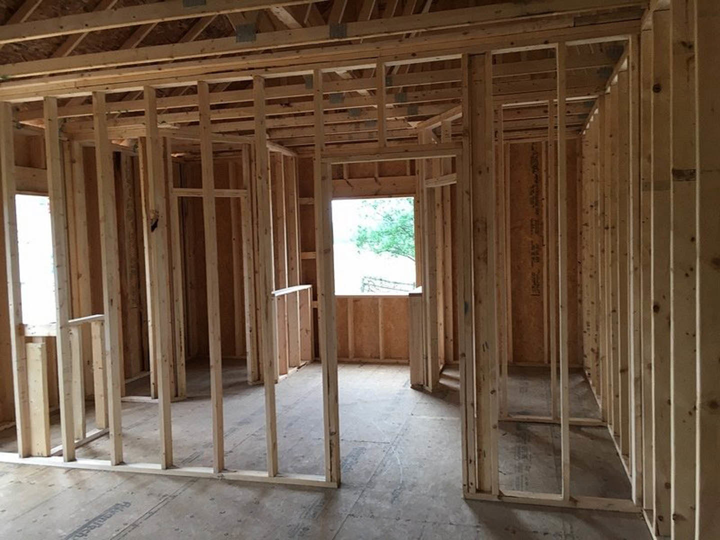 Room under construction with exposed wood framing, ceiling beams, and natural light streaming through a window with a tree visible outside