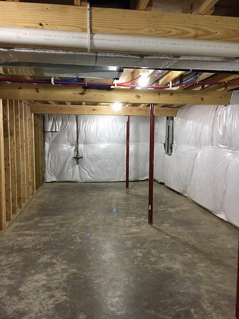 Basement with concrete floor, white plastic wall coverings, exposed wooden beams, white pipes, and a red support pole