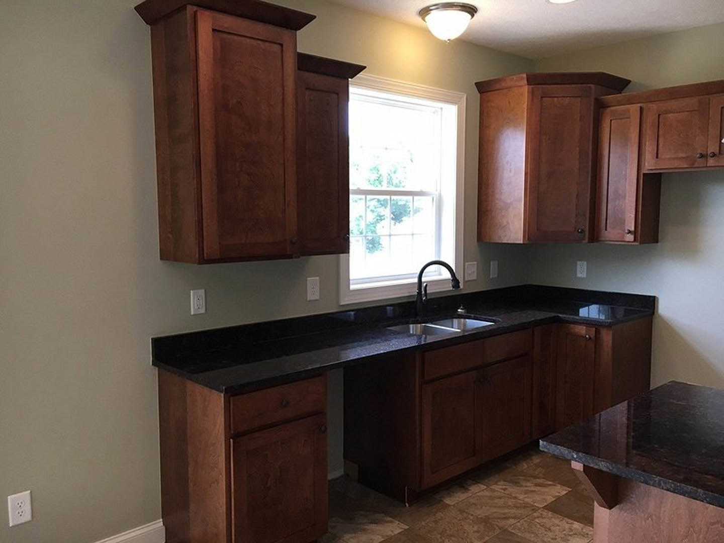 Dark wood kitchen cabinets, black marble countertop with built-in sink, white-framed window, modern light fixture, electrical outlet, and stainless steel kitchen appliances.