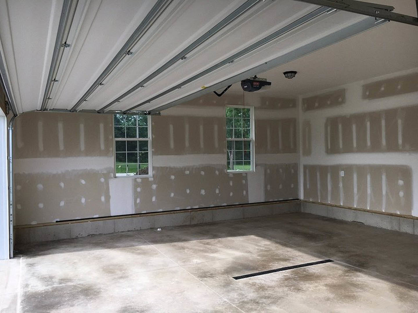 Empty room with white plaster walls, concrete floor, white-framed window, close-up of drain grate, and garage door opener.