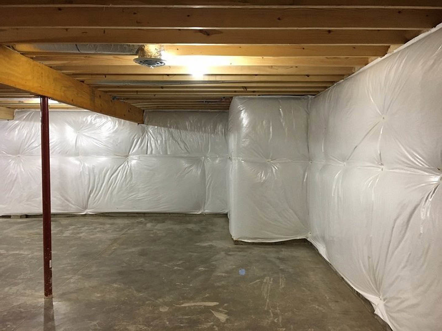 Basement room with exposed wooden beam, concrete floor covered in white plastic bags, plaster walls, ceiling light, and white leather upholstered bed