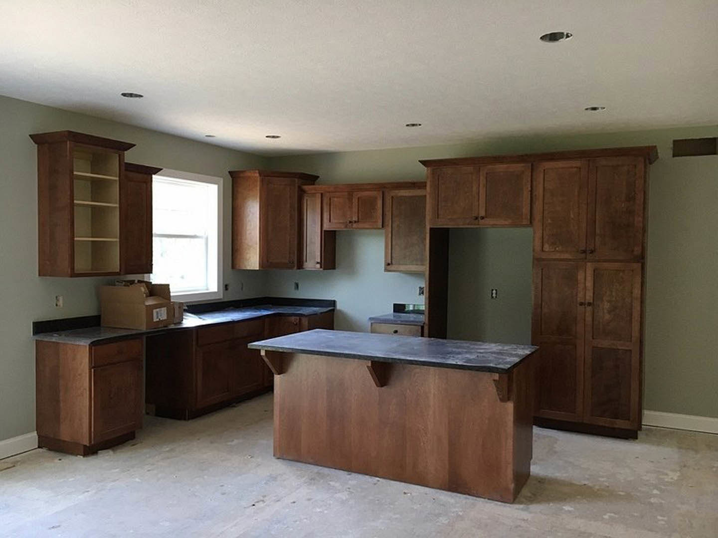 Kitchen featuring wood cabinets, black countertop, stainless steel sink, light streaming through window, cardboard box on floor, close-up of open wooden shelf and cabinet door