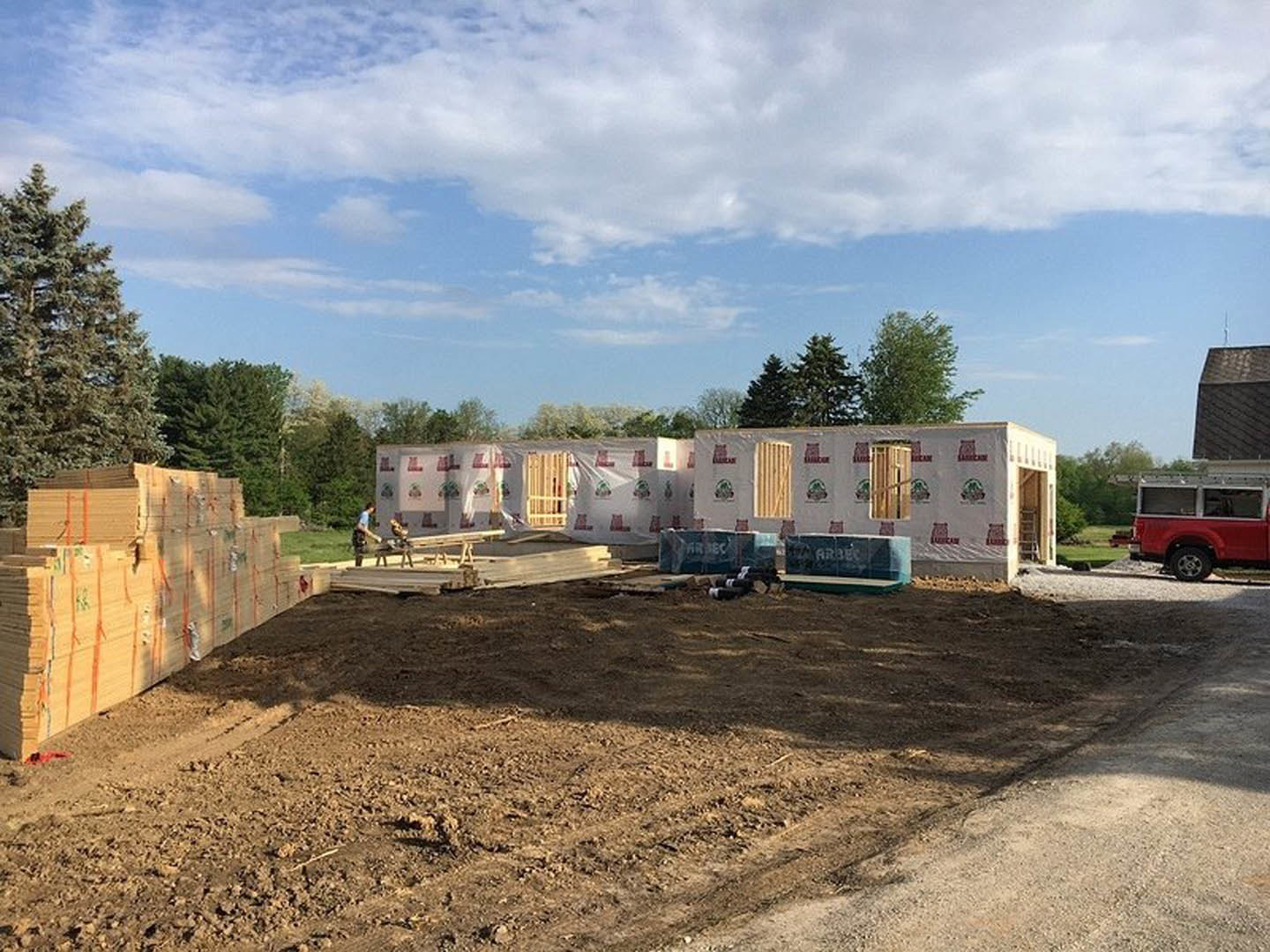 Partially built white house with protective covering, red truck parked nearby, large stack of lumber on dirt lot, scattered wood piles, overcast sky, trees in background