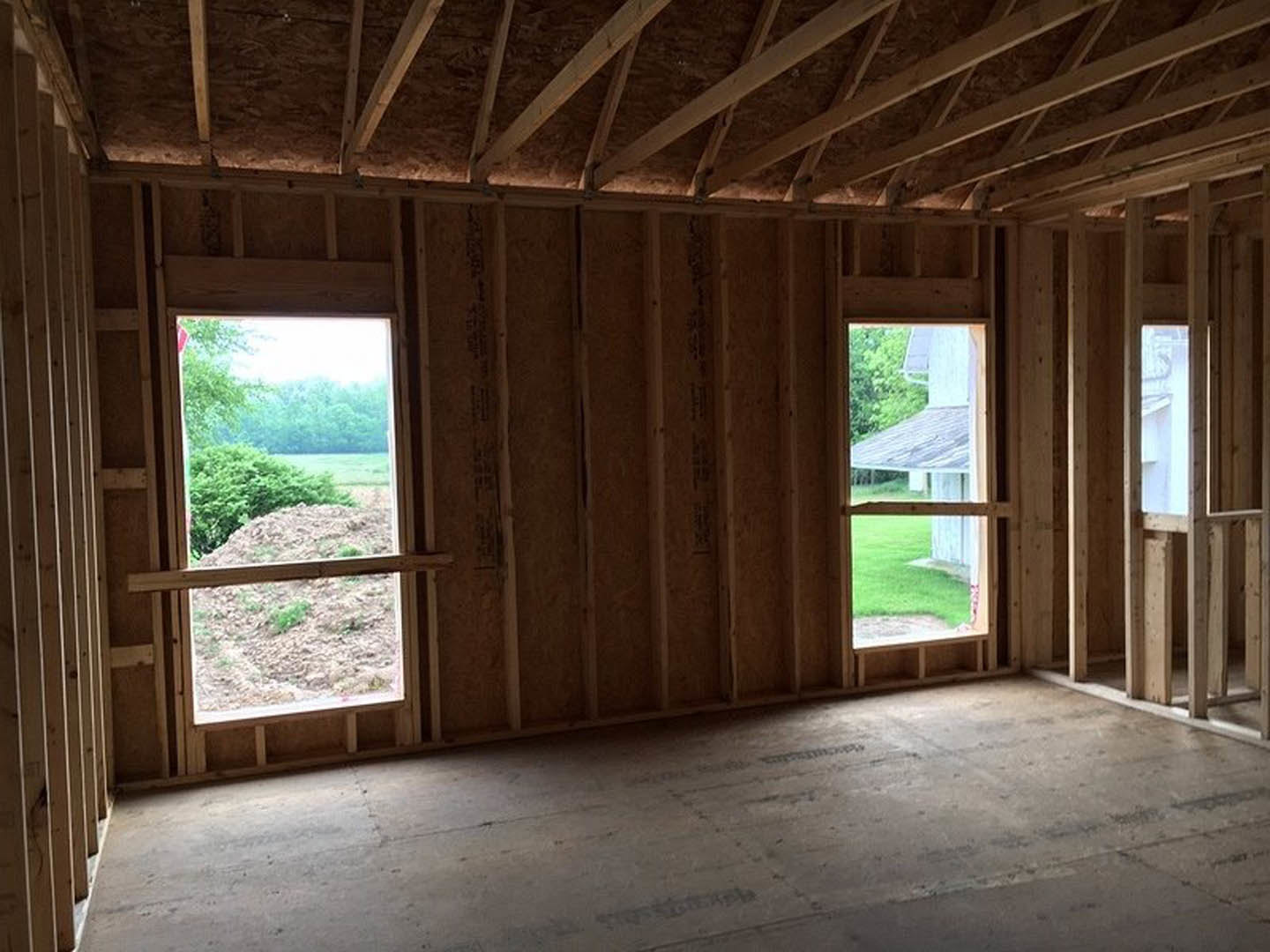 Spacious room featuring large windows, wood plank ceiling with exposed beams, hardwood flooring, and views of grassy yard and distant trees.