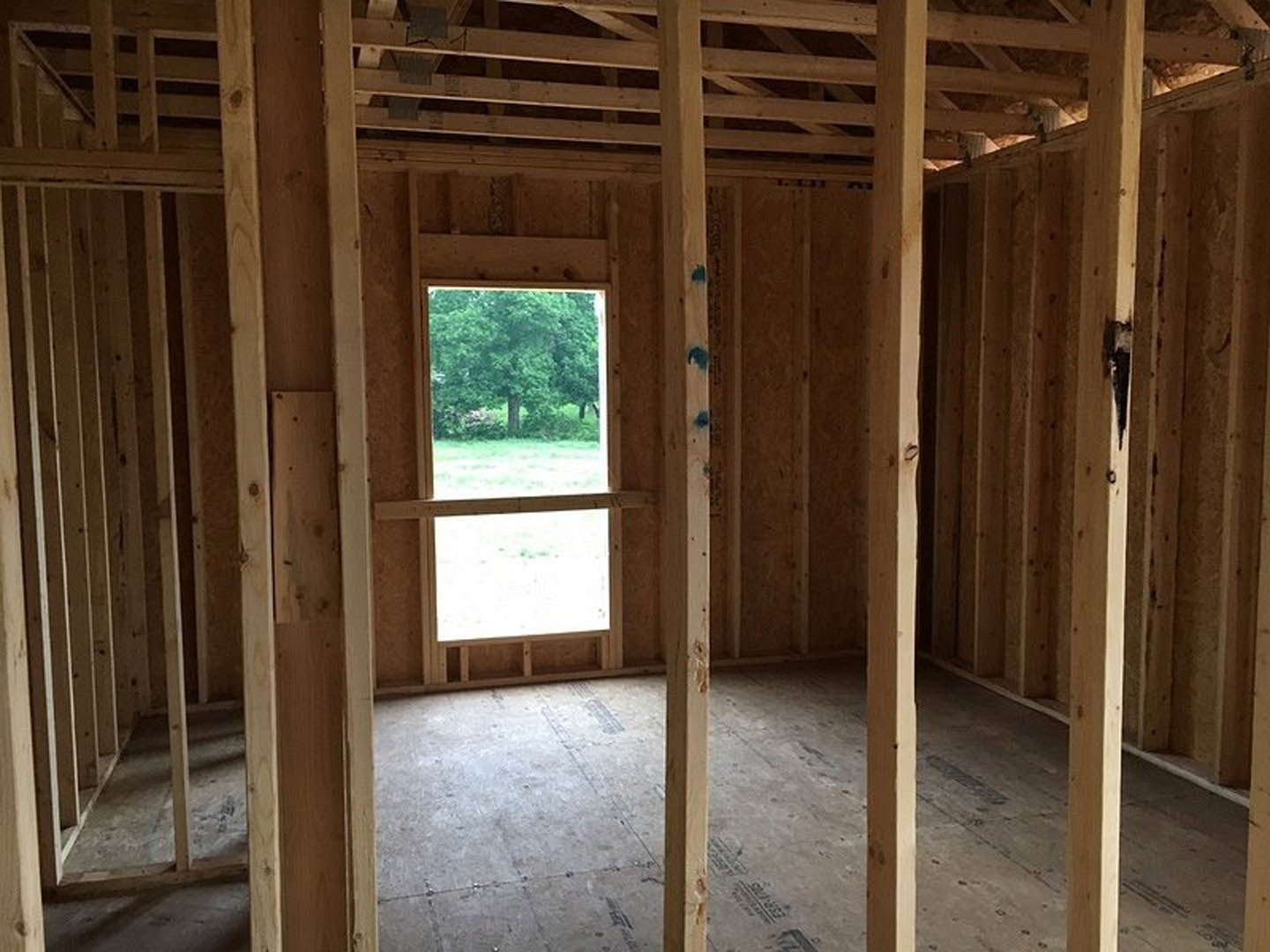 Room under construction with exposed wood framing, wooden slats on the wall, concrete floor, and large window framed in wood overlooking a tree outside.
