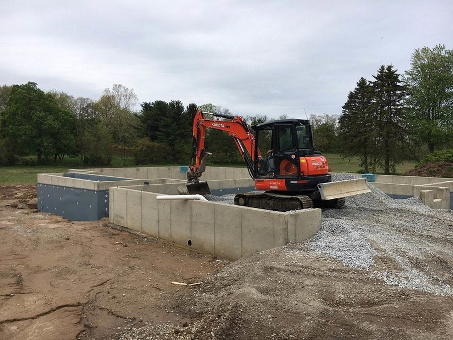 Bulldozer with open door parked on soil near concrete foundation wall and exposed pipe, surrounded by trees under cloudy sky