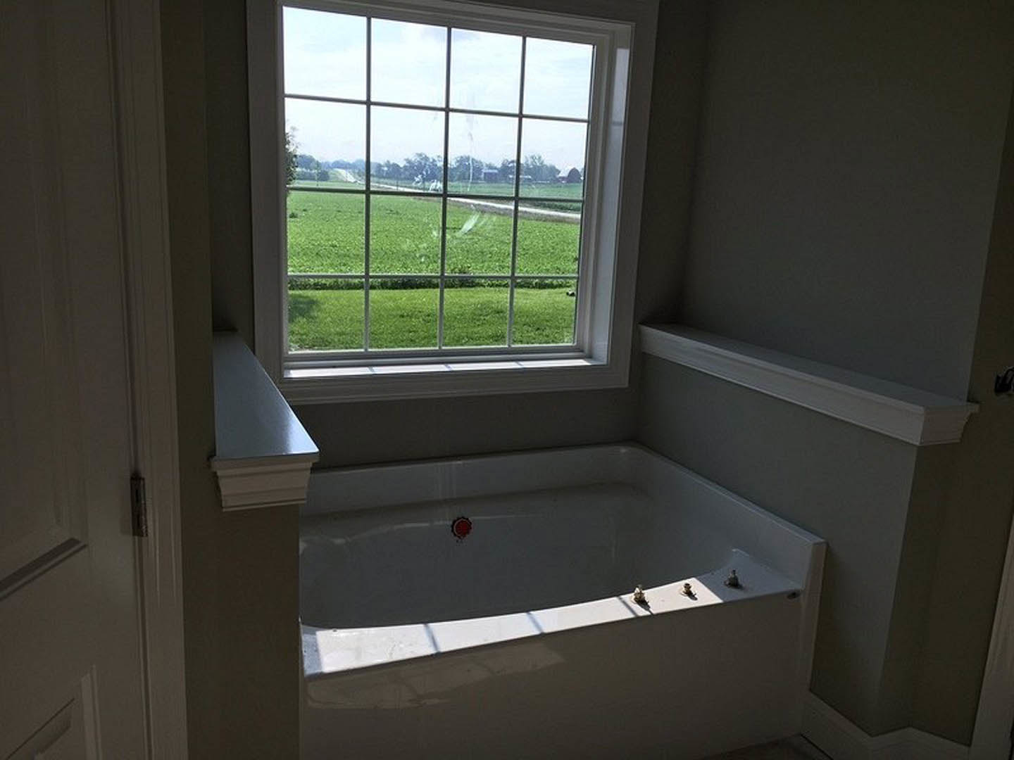 Freestanding white bathtub beneath large window overlooking grassy field, white countertop and molding, natural daylight fills bathroom
